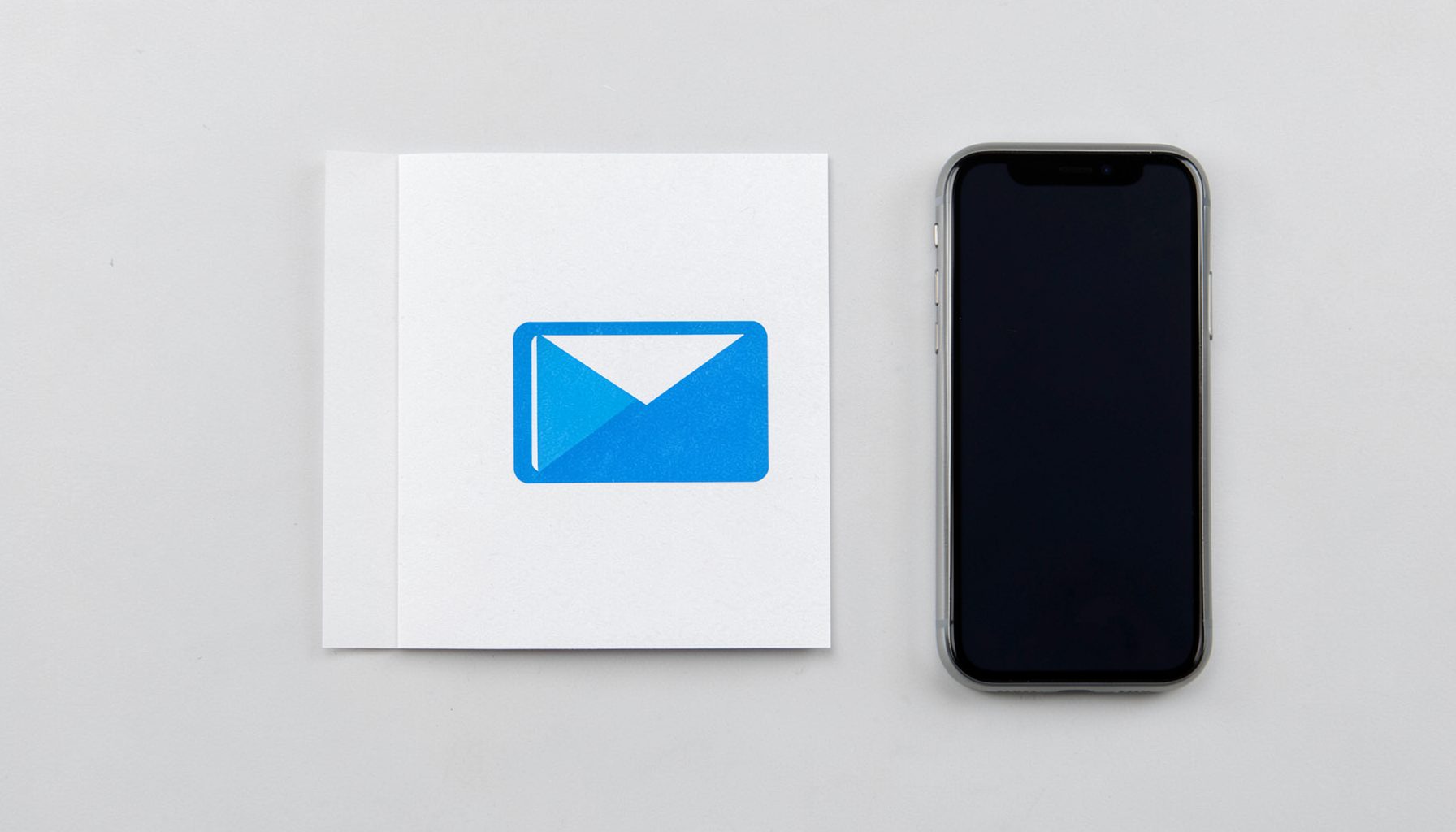 Flat-lay overhead photograph of an email icon printed on card stock, with a smartphone beside it on a pale gray background