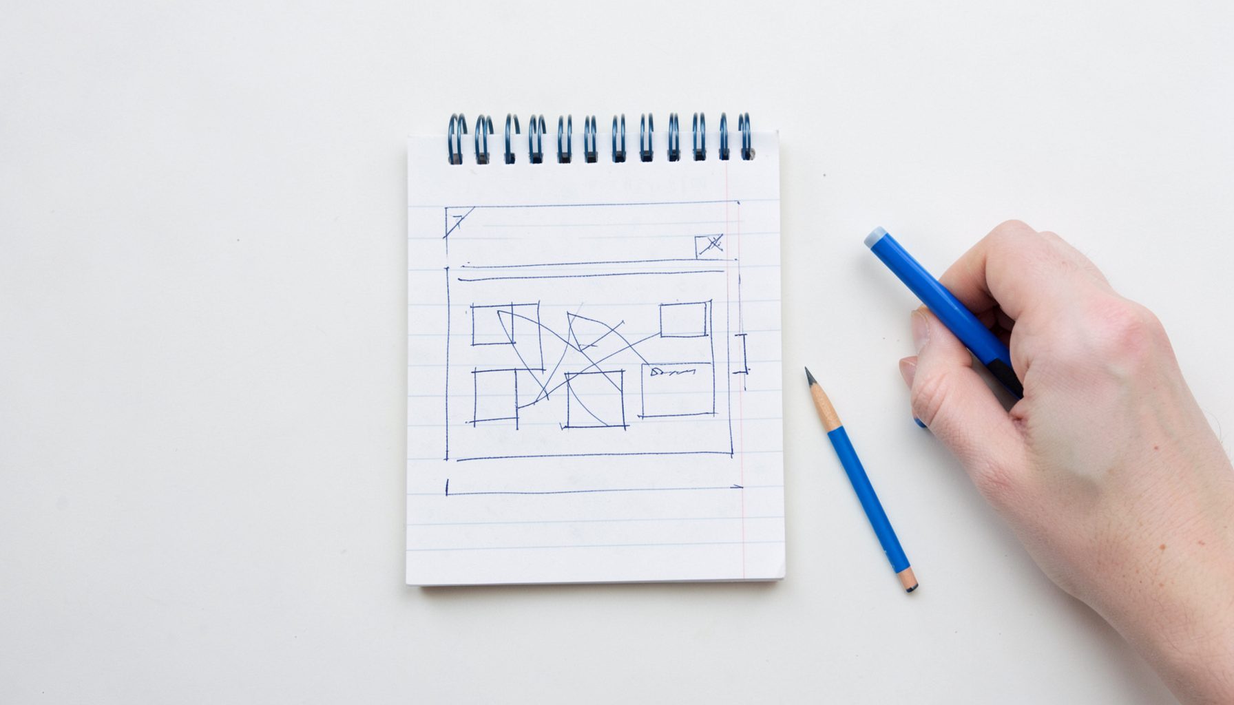 Flat-lay overhead photograph of a notepad with a hand-drawn website wireframe, pencil and eraser nearby, on a white surface